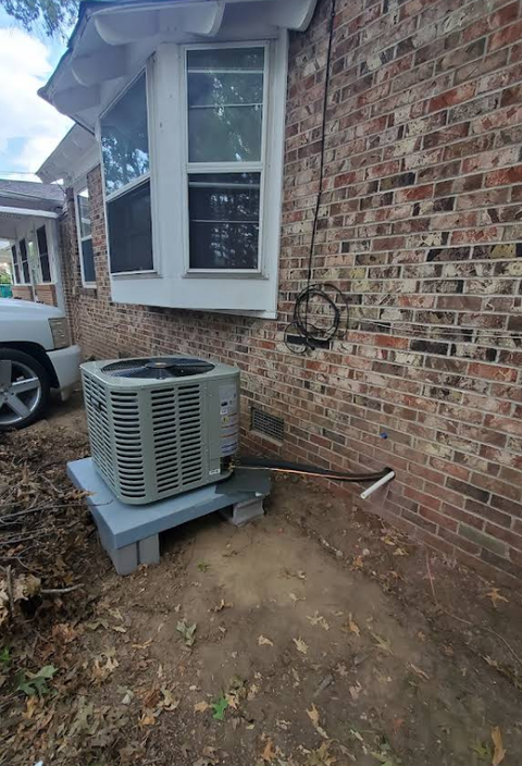 Exterior shot of an AC unit on concrete blocks next to a brick house with a bay window.