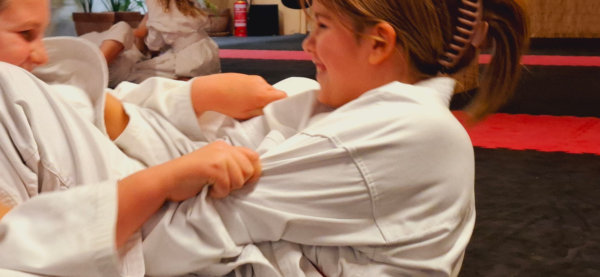 Zwei Mädchen kämpfen spielerisch am Boden und lachen, Partnerübung im Kinder-Kampfsport Thun.