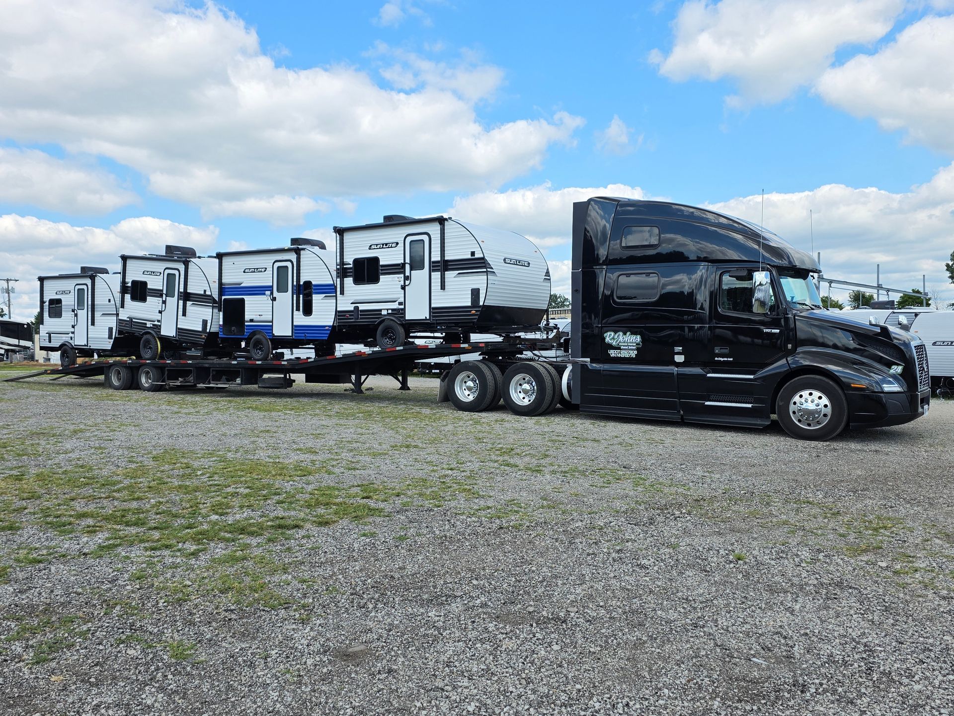 Black semi-truck hauling several recreational vehicles on a flatbed trailer under a cloudy sky.
