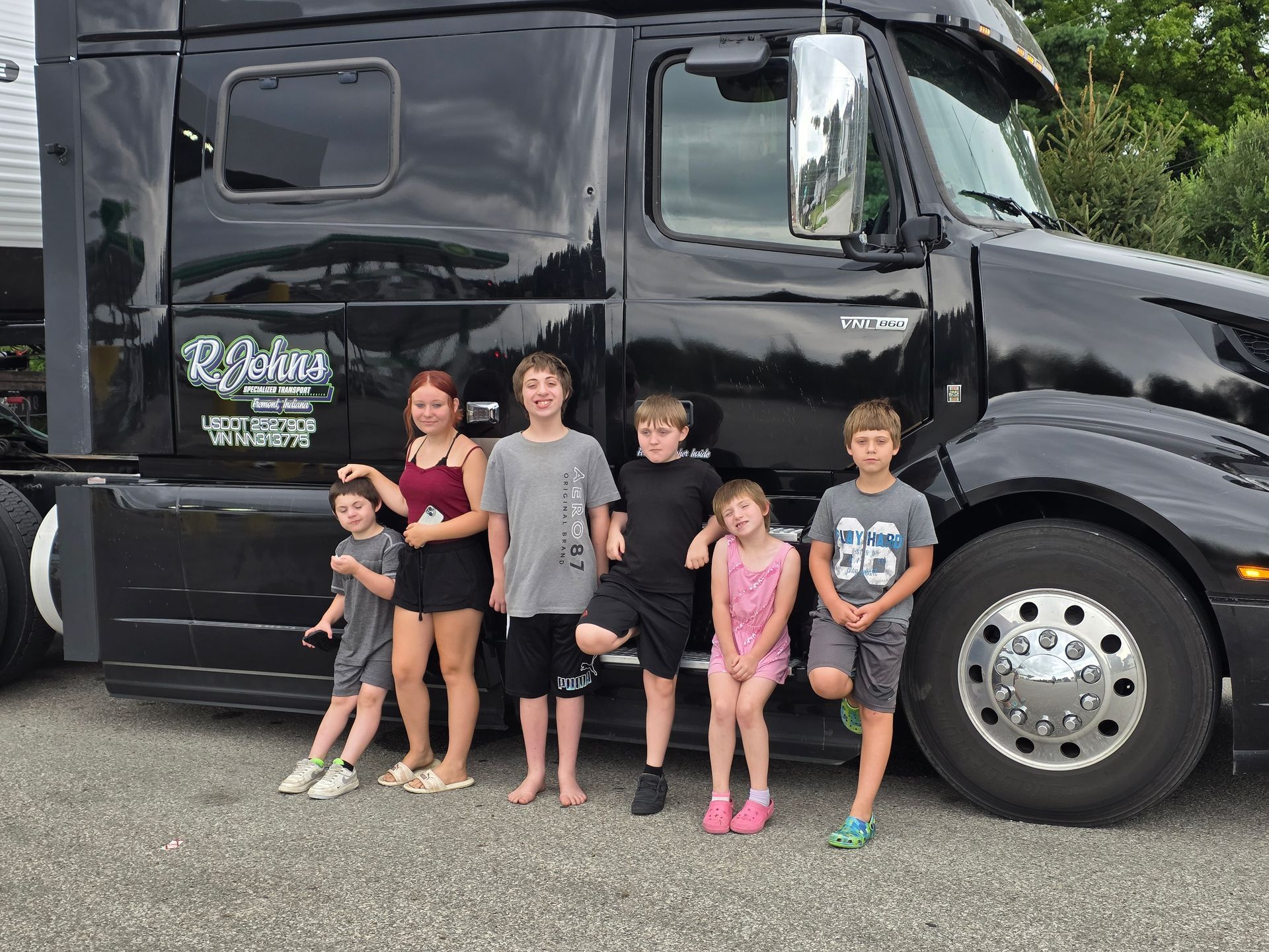 Group of kids pose in front of a black semi-truck. They stand on pavement, smiling.