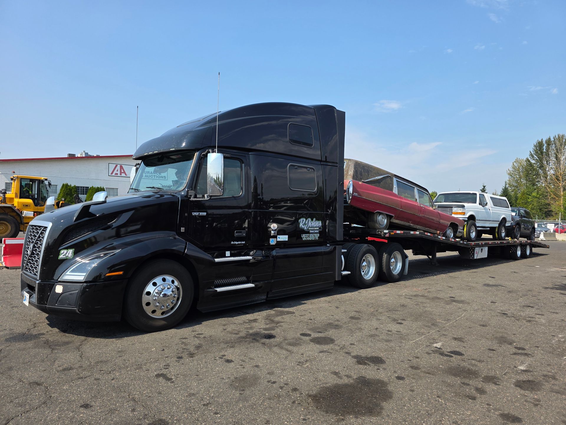Black semi-truck hauling a trailer with two vehicles on a paved lot on a sunny day.