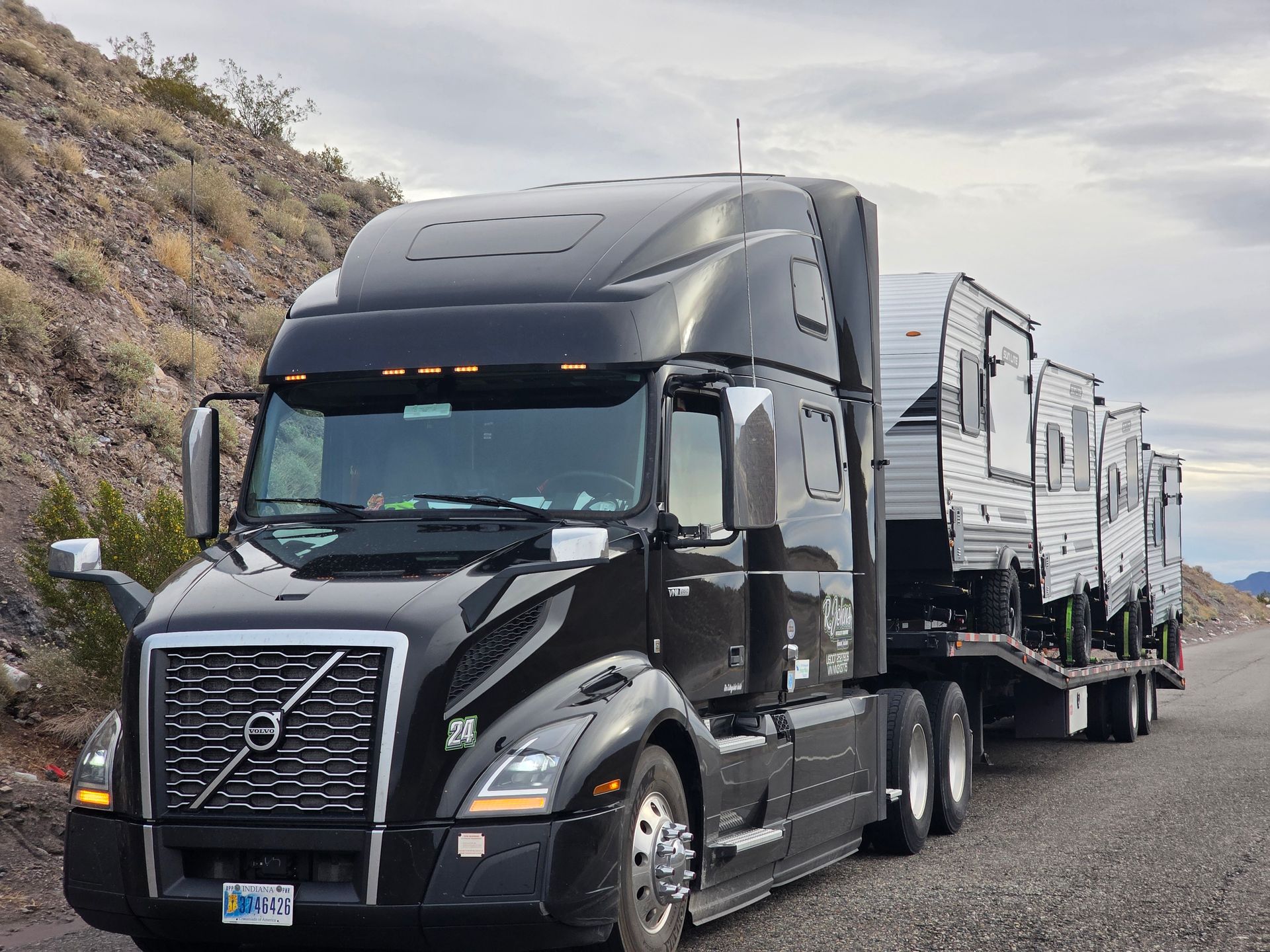 Black semi-truck hauling three travel trailers on a desert road with overcast skies.