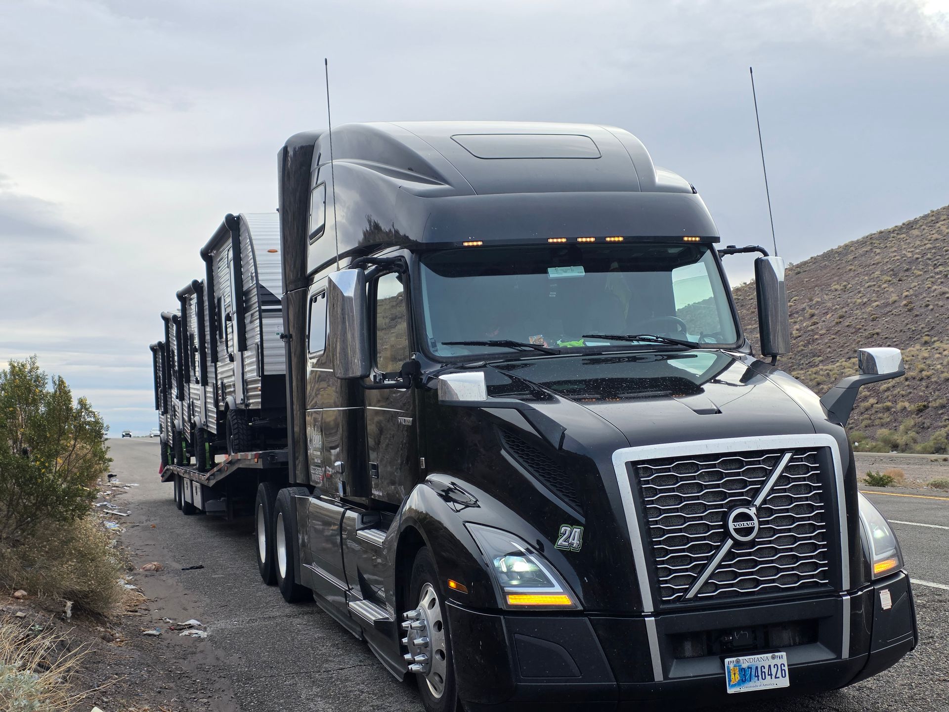 Black Volvo semi-truck pulling multiple trailers parked on a road. Cloudy sky, mountains visible in the background.