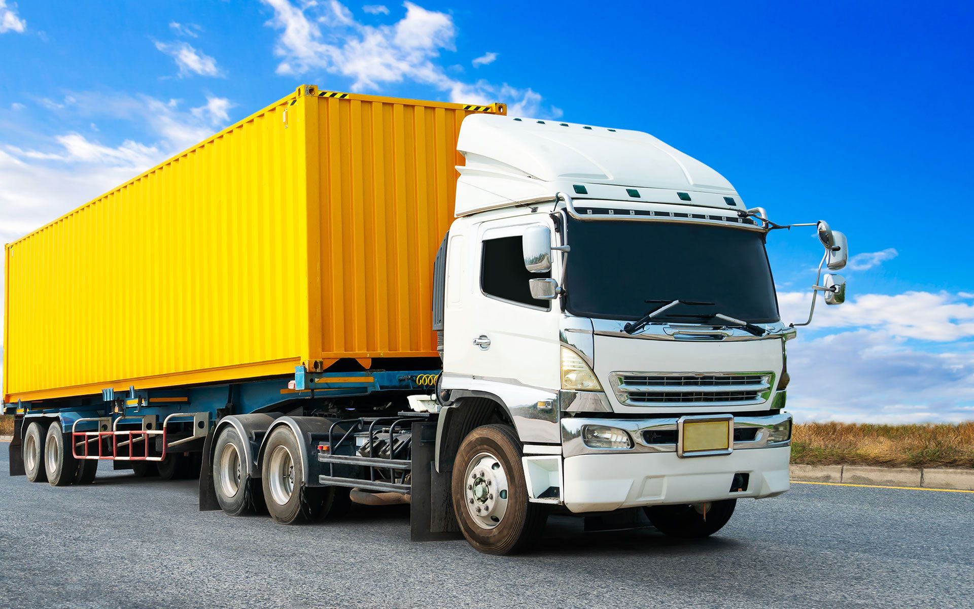 White semi-truck with yellow container on a road against a blue sky.