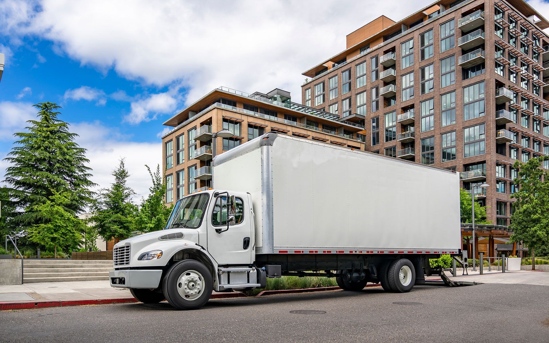 White box truck parked on a city street, tall apartment buildings in the background, blue sky with clouds.