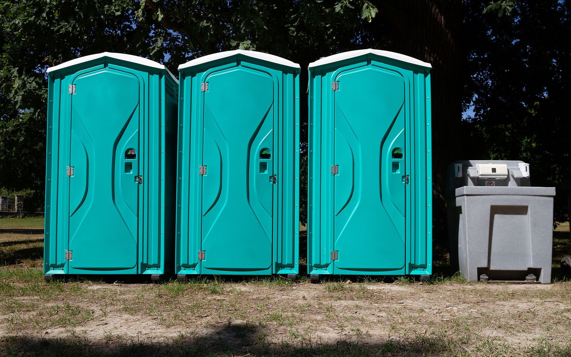 Three teal portable toilets and a gray handwashing station on grass outdoors.