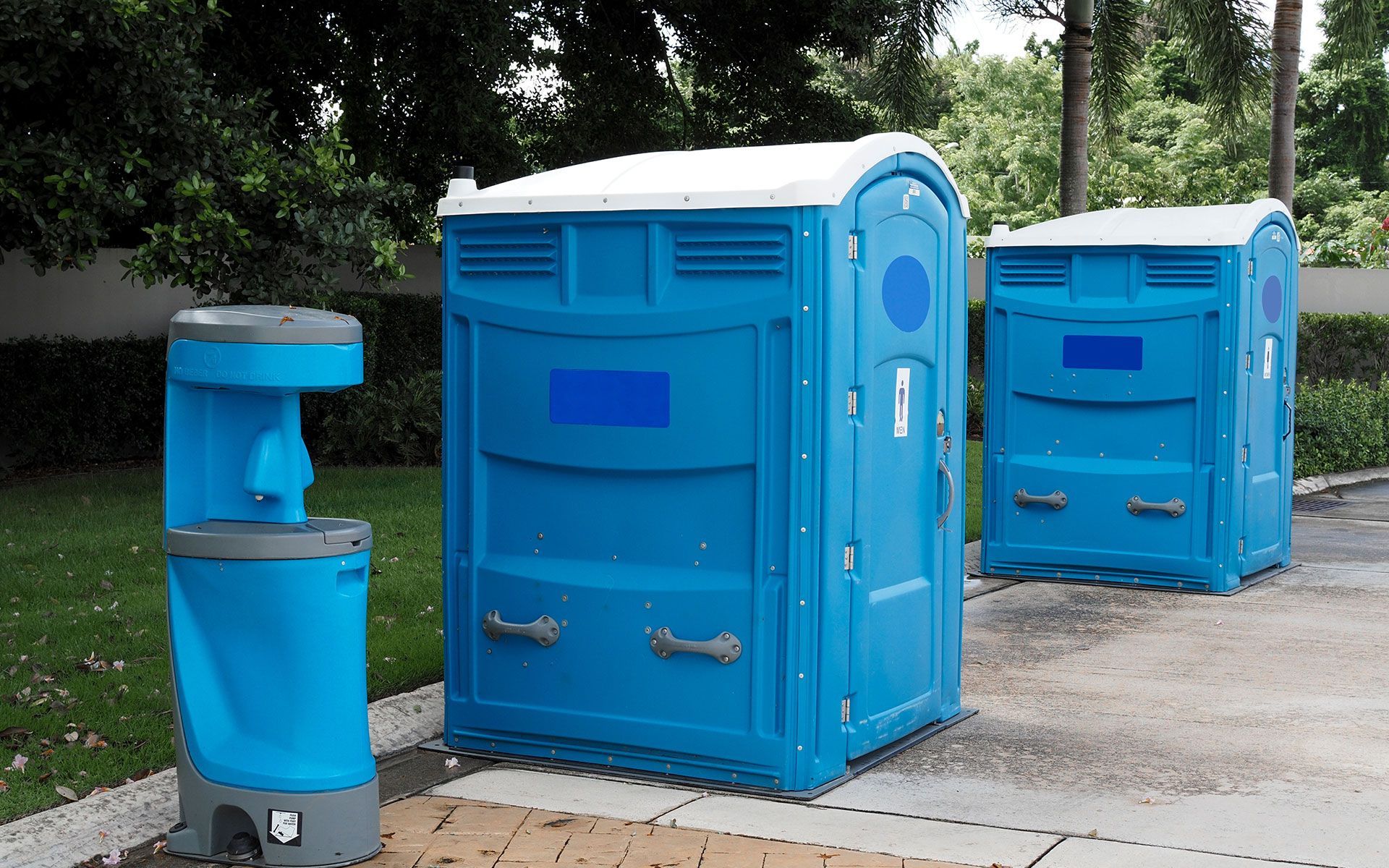 Blue portable toilets and a handwashing station on a paved area with trees in the background.
