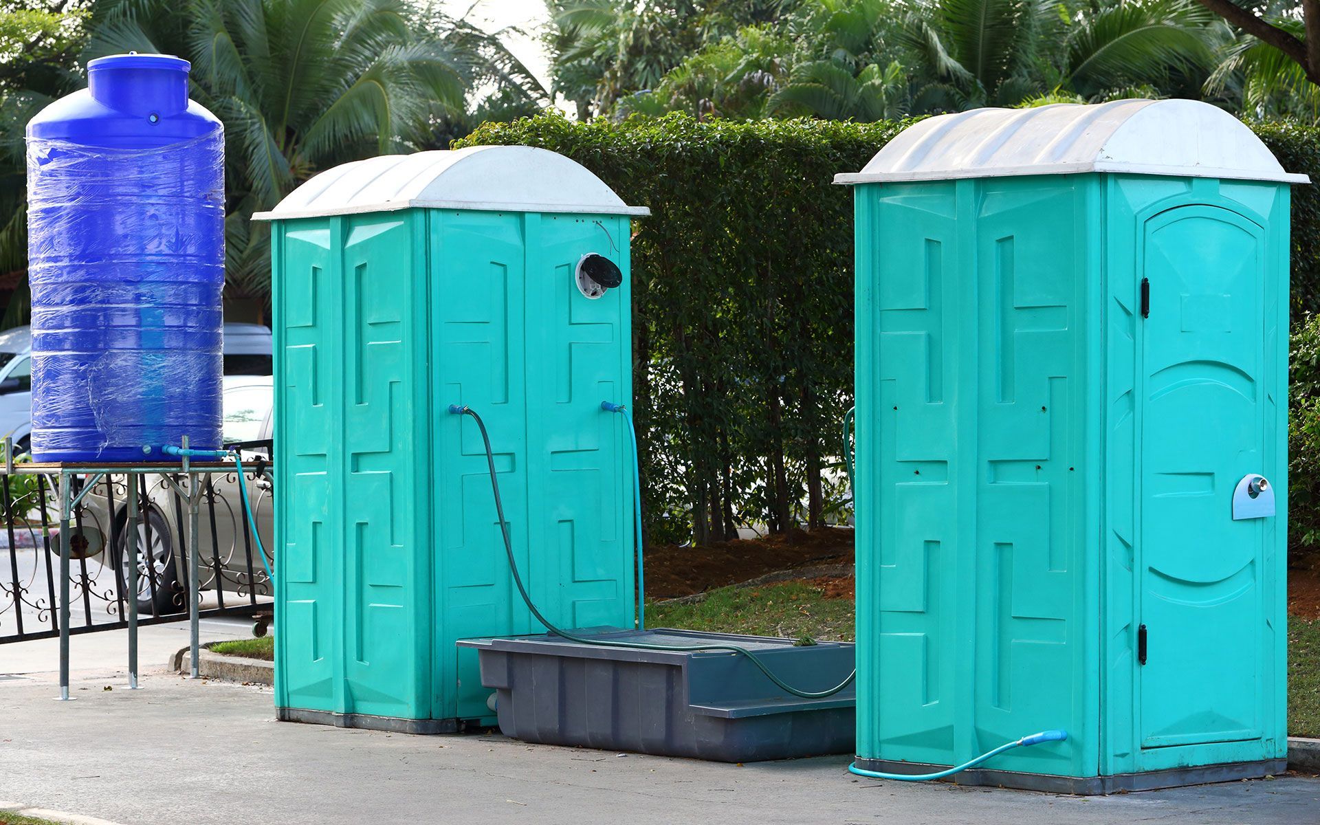 Two teal portable toilets with a blue water tank beside them on a paved area, next to greenery.