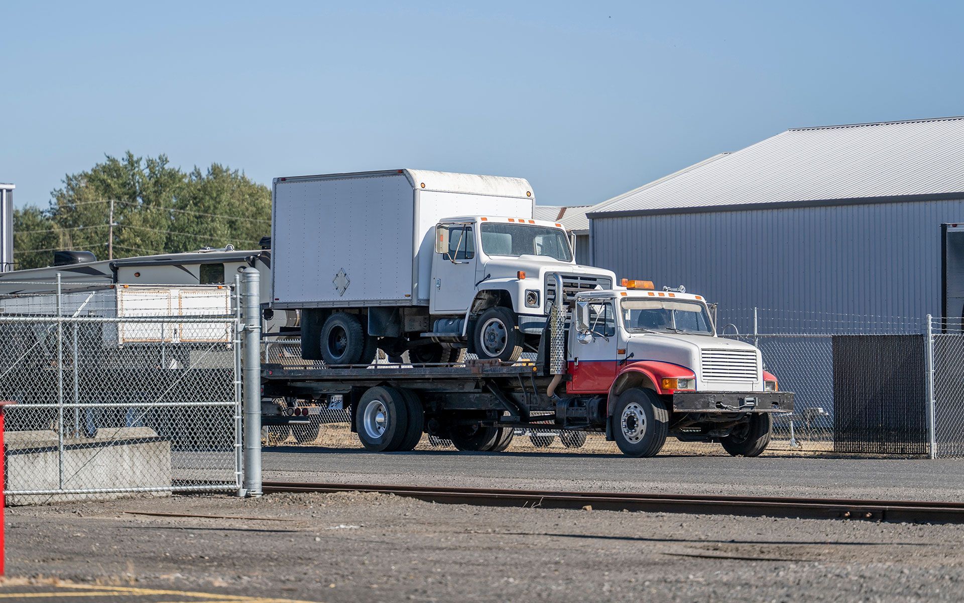 Truck hauling another truck with a white box in an industrial area.