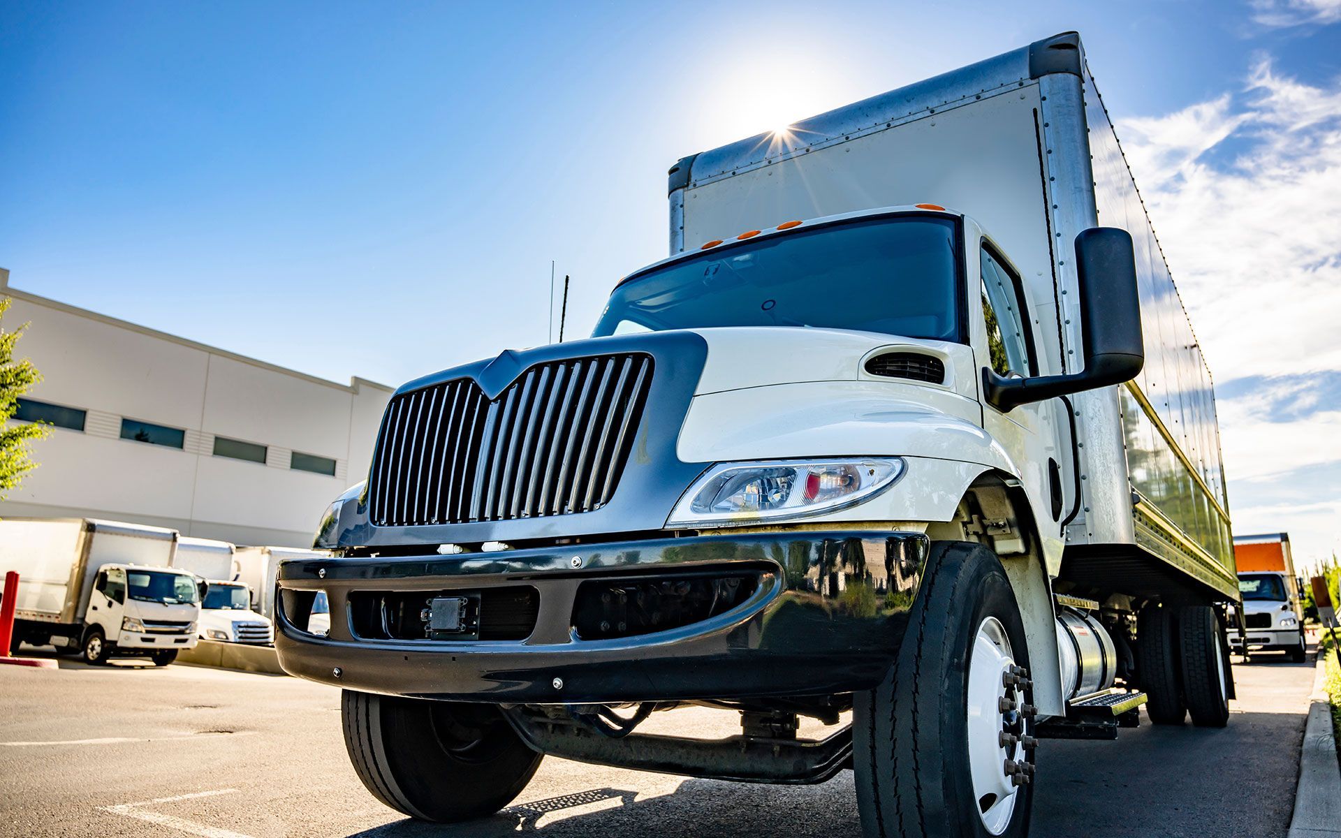 White semi-truck parked outdoors on a sunny day. Other trucks and a building are in the background.