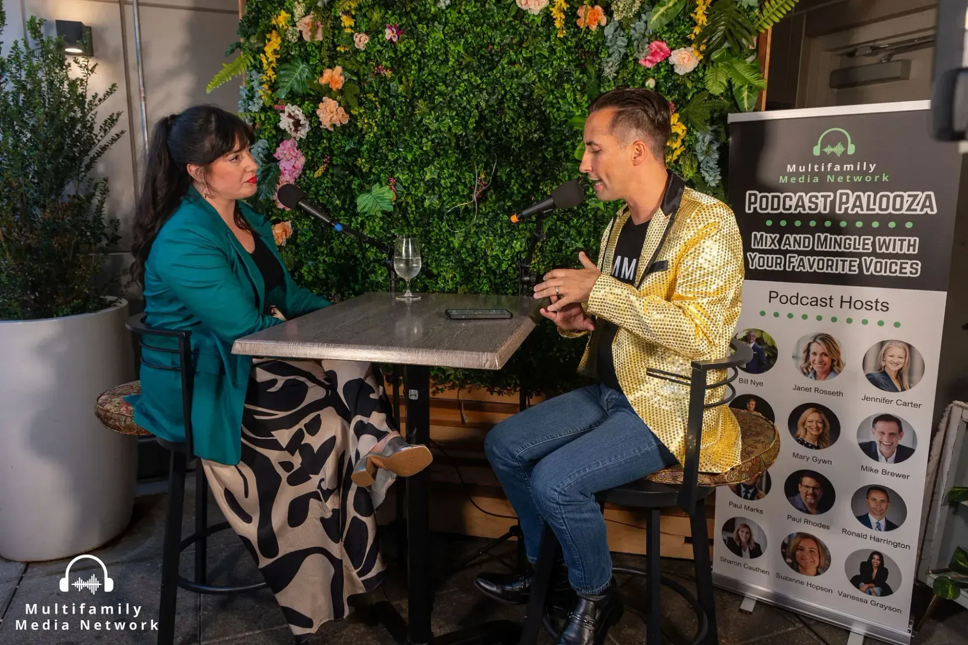 Man in a blue suit smiles, wearing glasses, pink tie, and pocket square, in front of greenery.