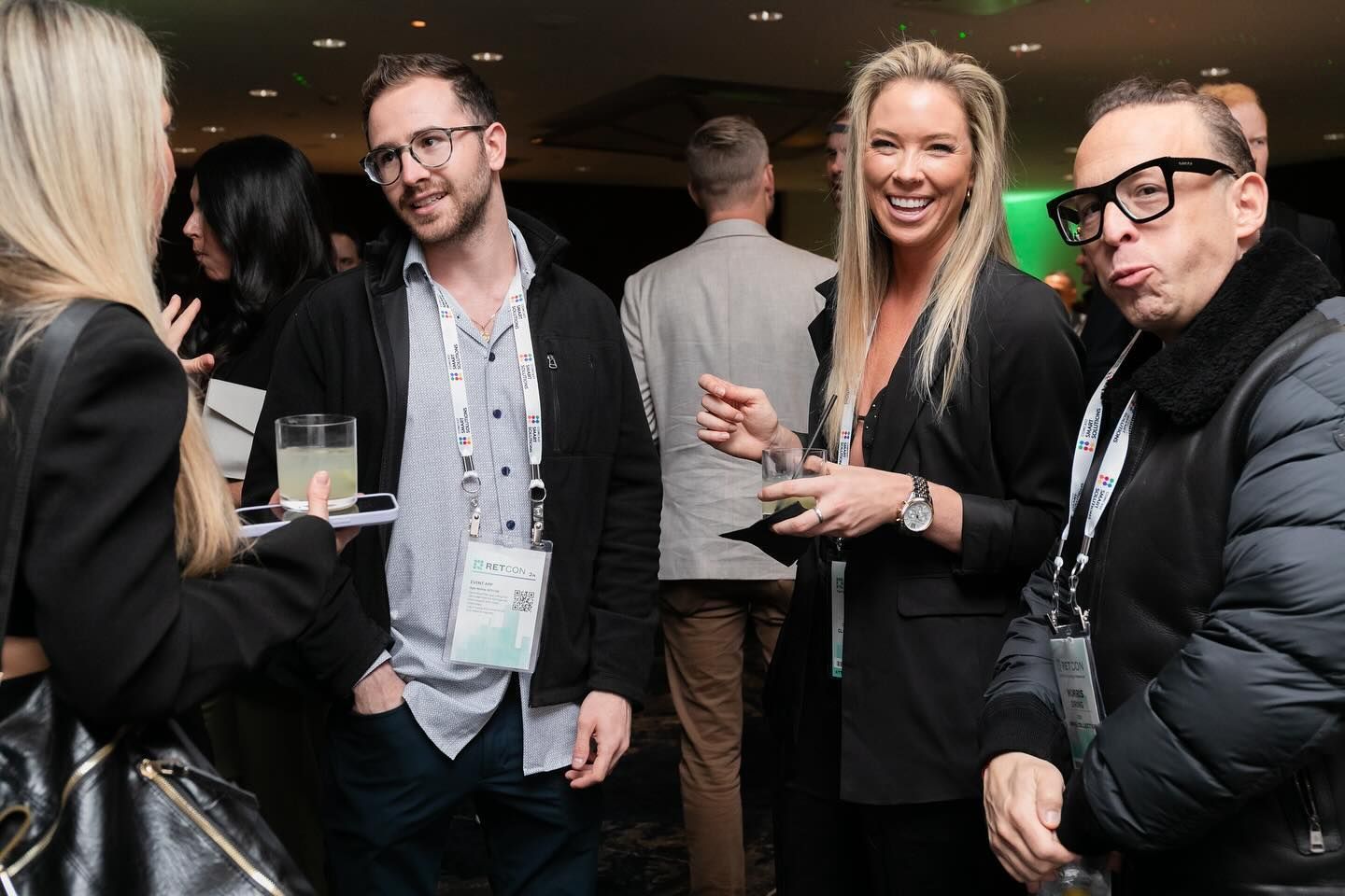 People socializing at an event, wearing lanyards. A woman with blonde hair smiles, a man makes a face.