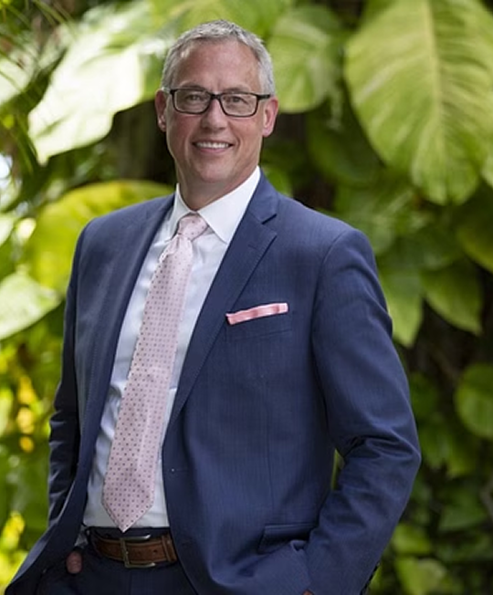 Man in blue suit, smiling, standing outside with green foliage in background.