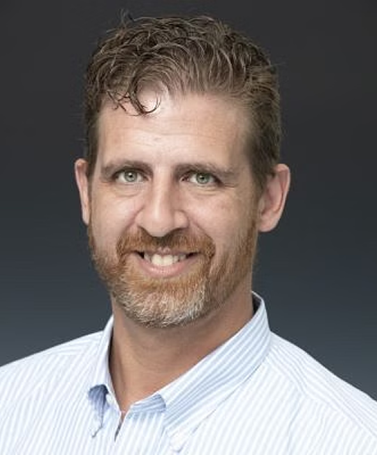 Man with brown hair and beard, wearing a light blue striped shirt, smiling.