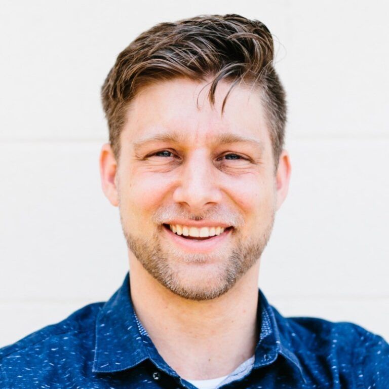 Man with short brown hair, small beard, smiling in front of a white wall, wearing a blue patterned shirt.