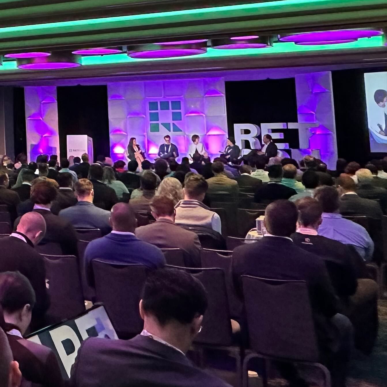 Panel discussion at a conference: speakers on stage, audience in seats, purple and green lighting.