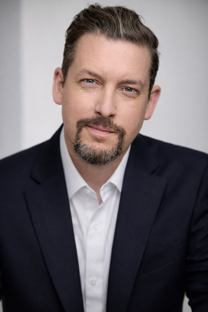 Man in a navy suit and white shirt, with a trimmed beard, looking at the viewer.