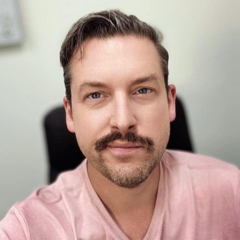 Man with short dark hair, mustache, and goatee, wearing a pink shirt, smiling.