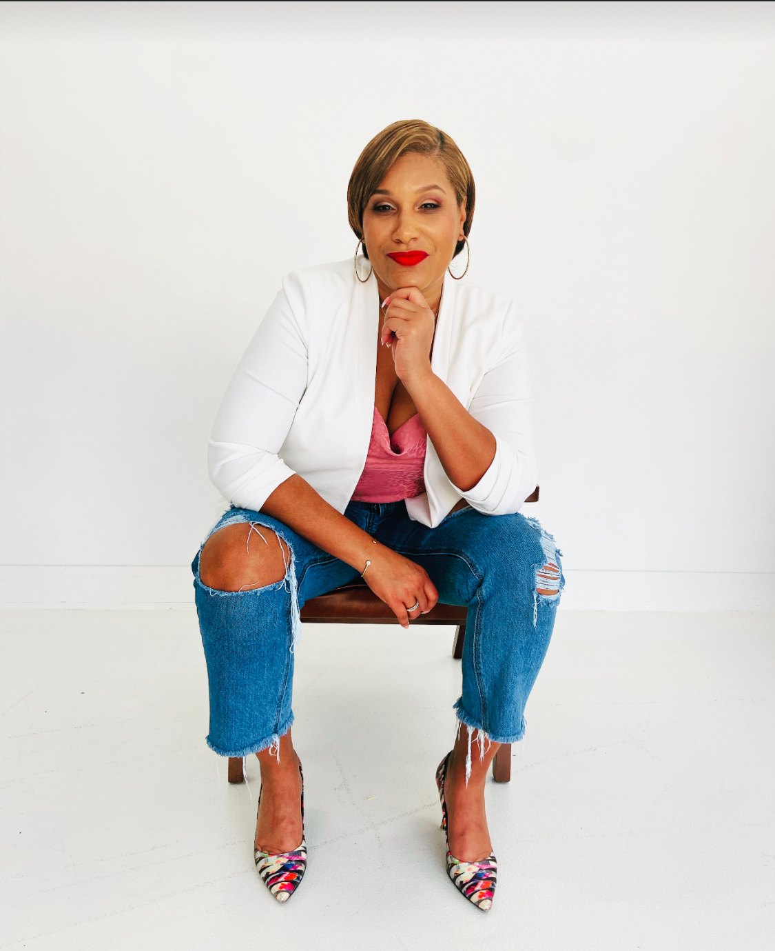 Woman in ripped jeans, blazer, and heels, seated indoors, hand on chin, bright red lipstick.