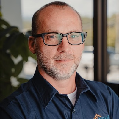 Man with glasses and a beard in a blue shirt smiles.