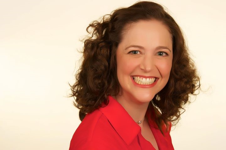 Woman with curly brown hair smiles brightly, wearing a red top, against a pale yellow background.