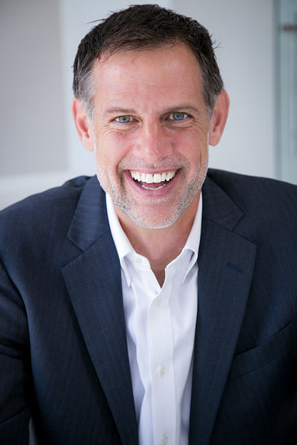 Man in a navy blazer and white shirt smiles broadly, indoors.