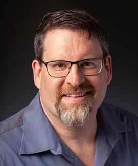 Man with glasses, beard, blue shirt, smiling at camera against a dark background.