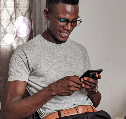 Man in glasses smiling, using a phone. Leaning against a wall, neutral background.
