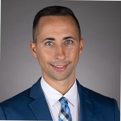 Man in blue suit, white shirt, and patterned tie, smiling. Gray background.