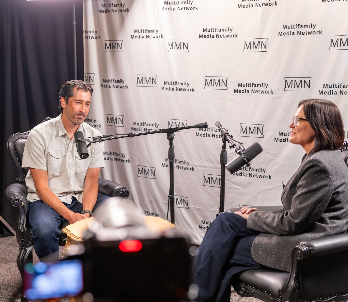 Person with glasses and headphones holding a microphone, looking to the side. Wearing a blue sweatshirt