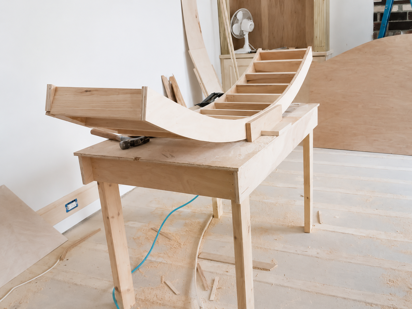 Wooden sailboat model on a workbench in a workshop, with unfinished planks and tools nearby