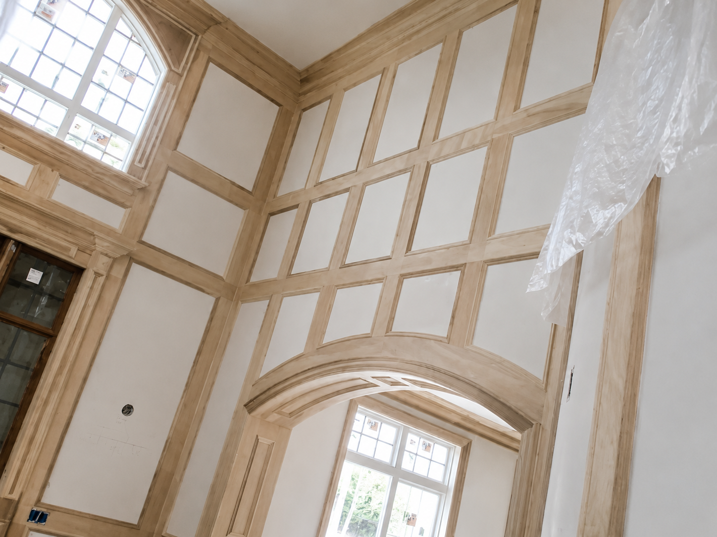 Tall interior wall with cream wood paneling, arched window, and scaffolding or plastic sheeting at right.