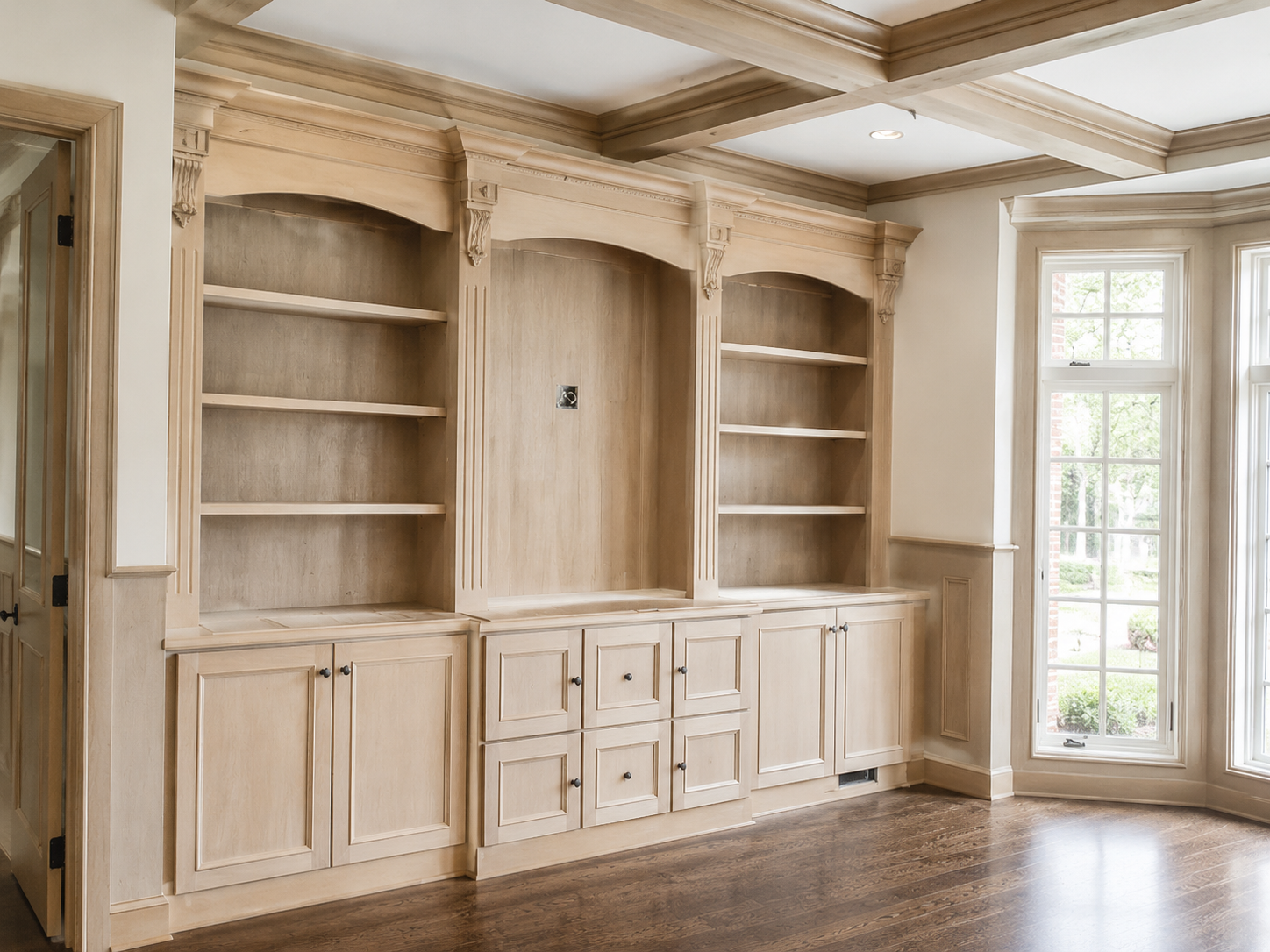 Built-in wooden cabinets and shelves in a bright, empty room with a coffered ceiling and tall windows