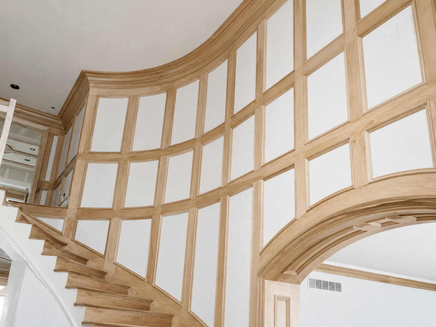 Curved wooden staircase with white paneled wall and arched opening in a bright interior