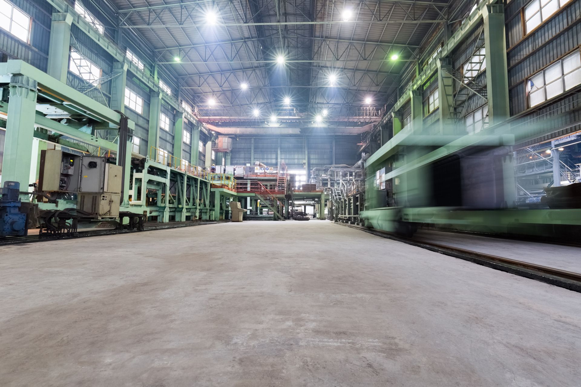 Inside of a factory, green machinery, concrete floor, overhead lighting, and a blurred green train on a track.