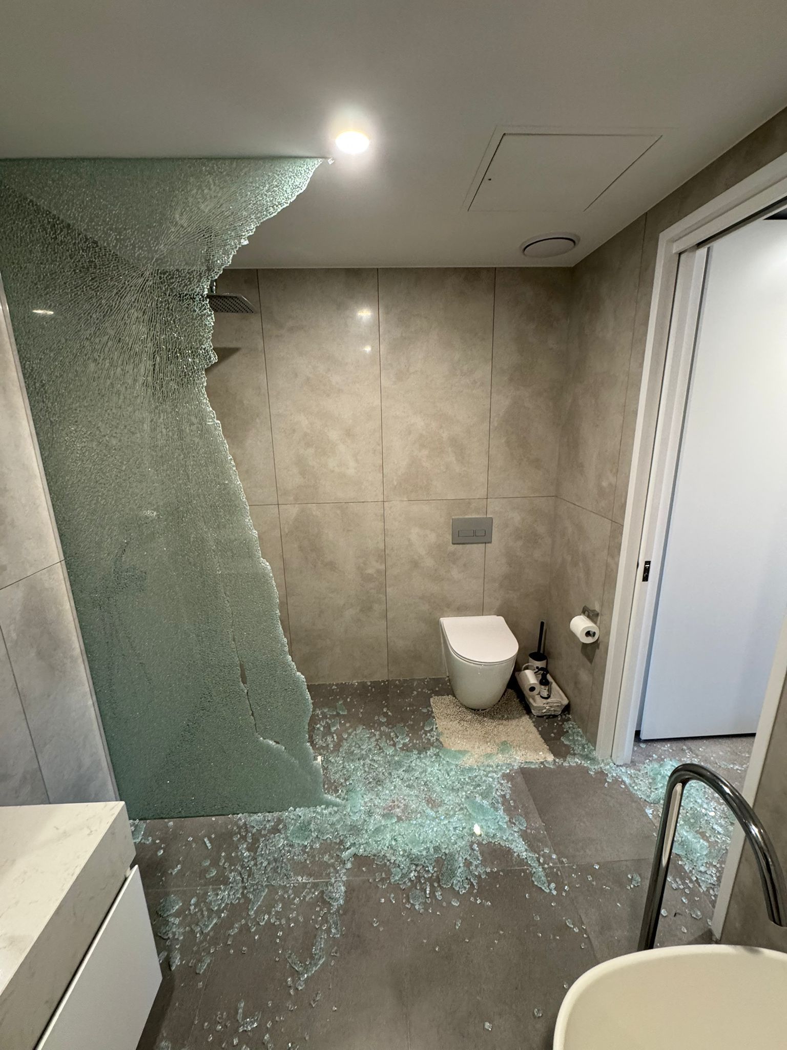 Bathroom with shattered glass shower wall, shards on floor, toilet, and open door.