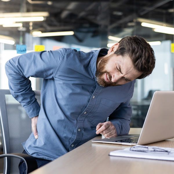 A man in a blue shirt hunched over a laptop at an office desk, clutching his lower back in apparent pain.