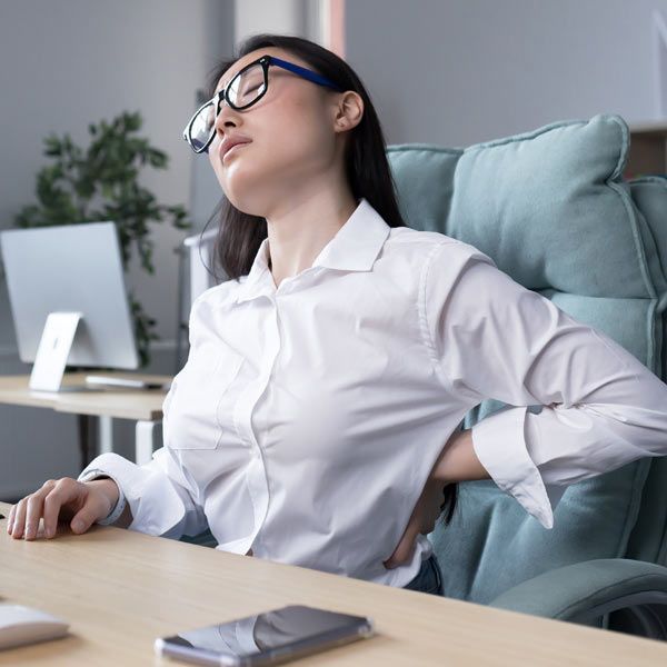 A person with poor posture in a white shirt sits at an office desk, leaning back with their eyes closed, holding their lower back in pain.