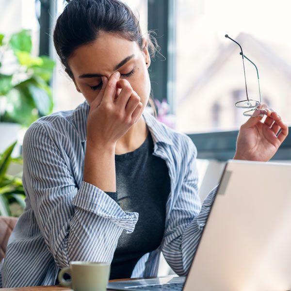 A person sitting at a desk with a laptop and a coffee cup, looking stressed while rubbing the bridge of their nose.