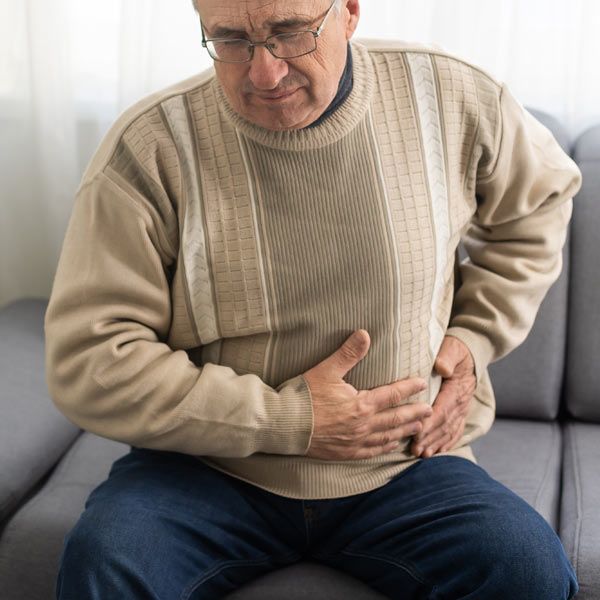 A man with a Hernia wearing a beige sweater and glasses, sitting on a sofa and holding their abdomen while showing signs of pain.