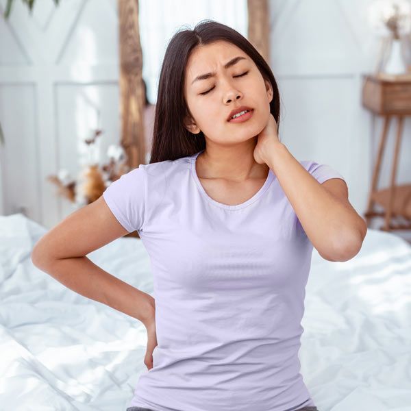 A person in a light purple shirt sitting on a bed, holding their neck and lower back while grimacing in pain.