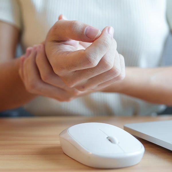 A person with Carpal Tunnel holding their wrist in pain while sitting at a desk with a white computer mouse and a laptop.