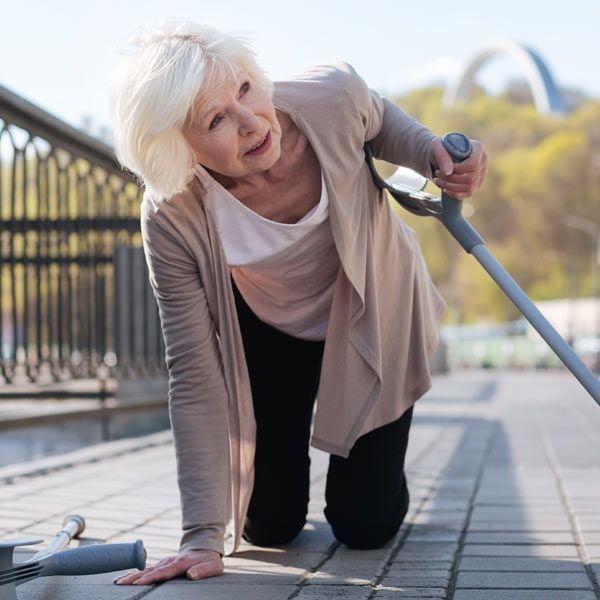 A woman who lost her balance is on their knees on a paved path, holding one crutch with another lying on the ground.