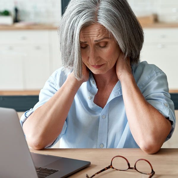 A woman with Fibromyalgia with gray hair sits at a desk with a laptop, looking down and holding the back of their neck in discomfort.