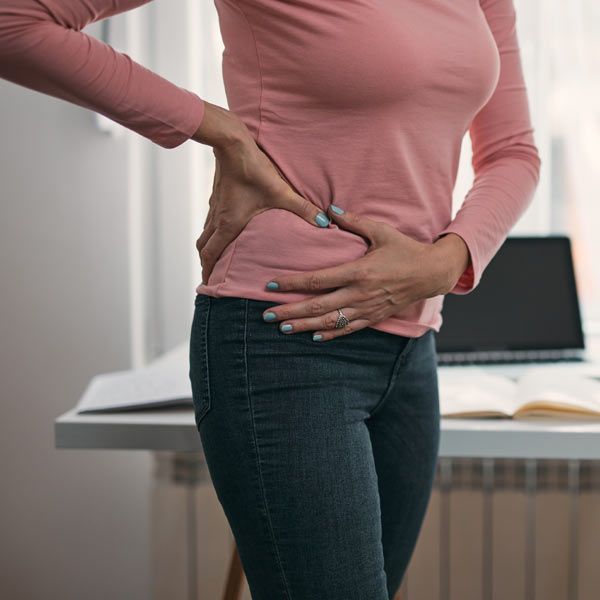 A person with hip pain in a pink shirt and jeans stands at a desk, holding their side while appearing to experience pain.