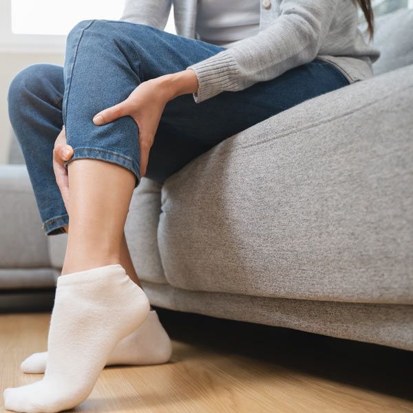 A woman with Muscle Cramps is sitting on a gray couch, holding their lower leg, which suggests pain or discomfort.