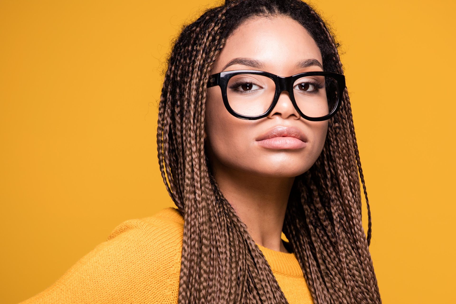Woman with braided hair wearing large black glasses, yellow sweater against a yellow background.