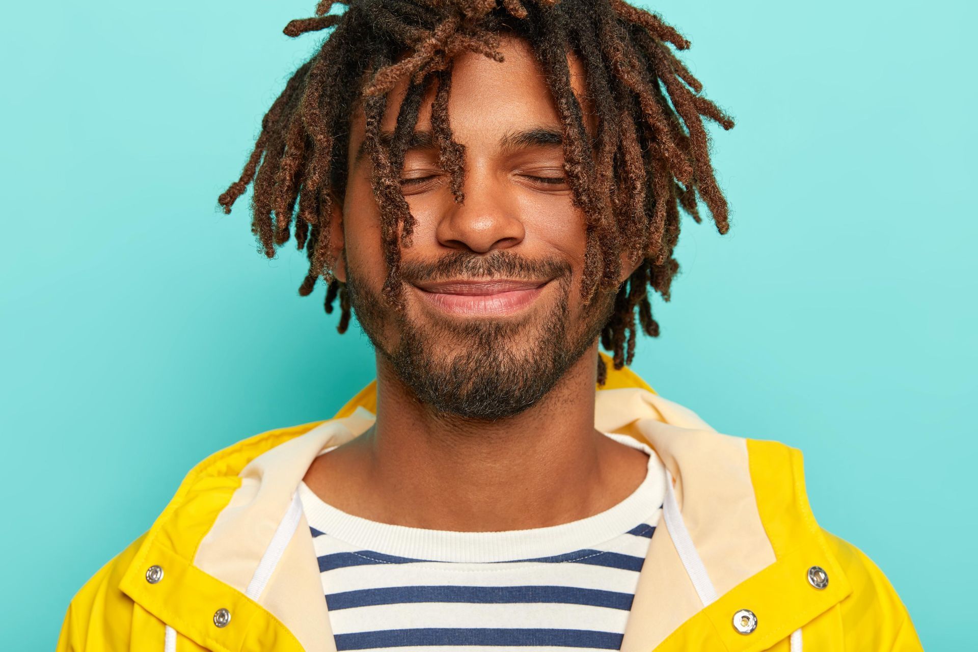 Man with dreadlocks and closed eyes, wearing a yellow jacket and striped shirt. Light blue background.