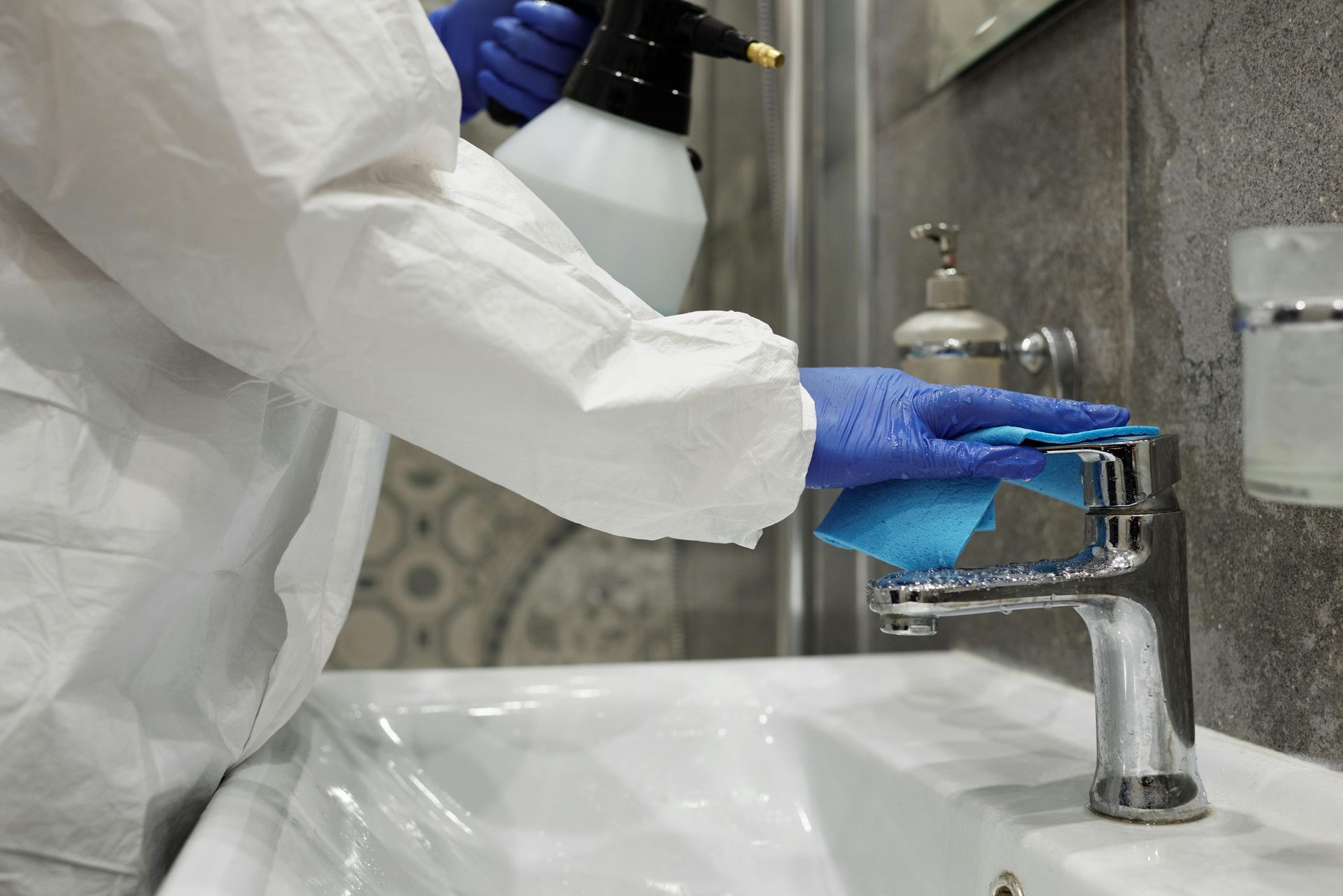 Person in protective suit cleaning a bathroom sink with spray and a cloth.