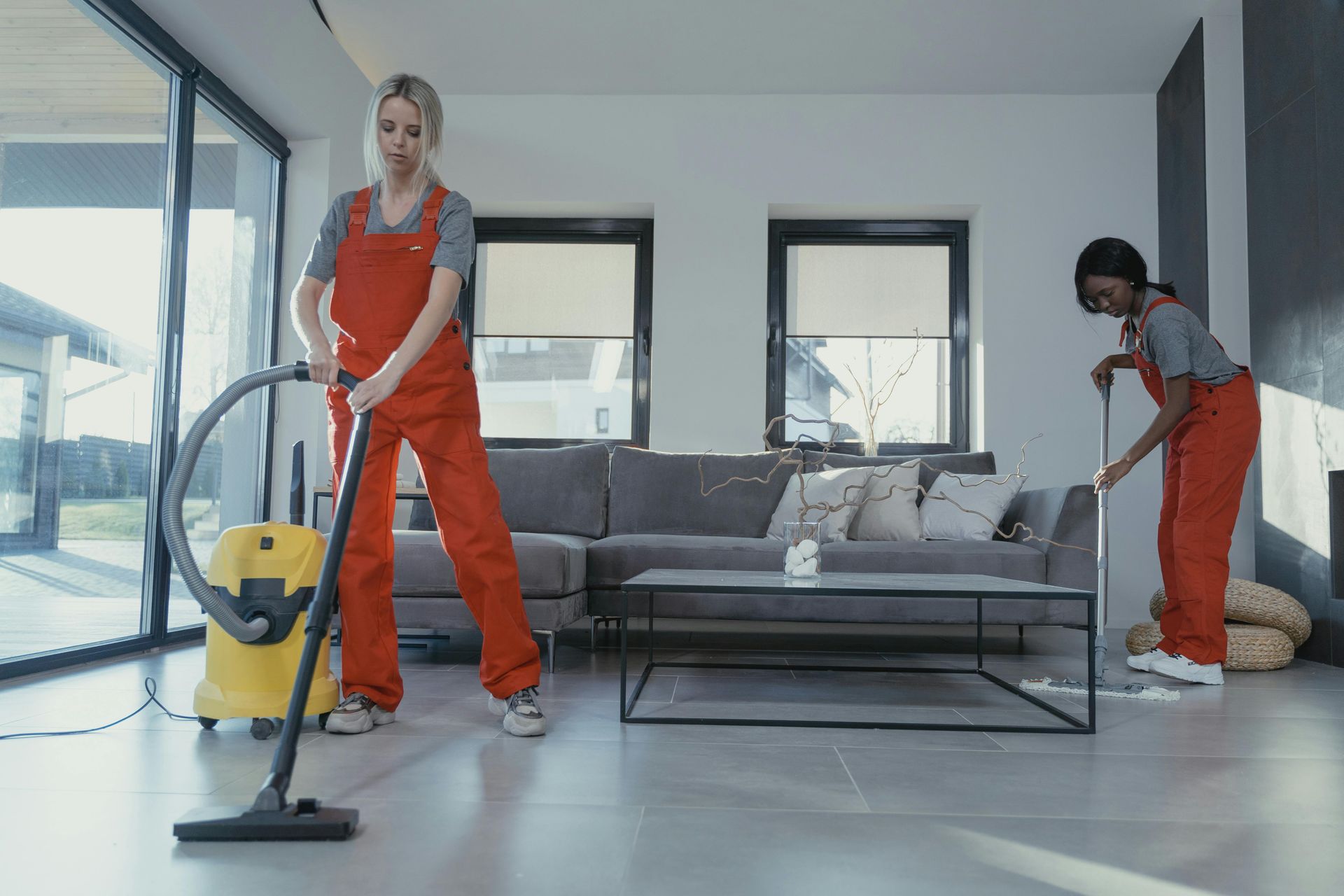 Two women in orange jumpsuits cleaning a modern living room. One vacuums, the other mops.